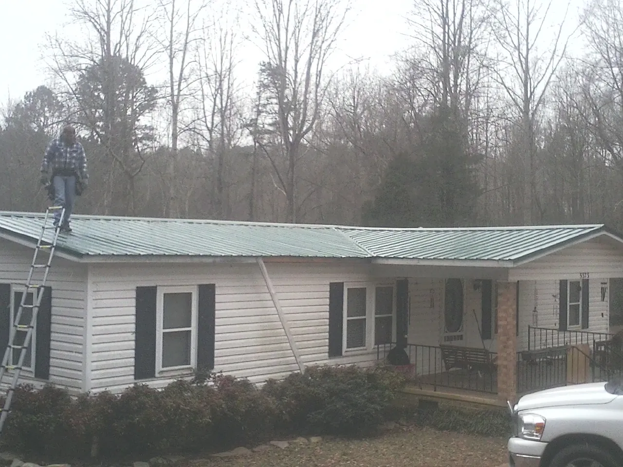 Worker on a ladder beside a new green metal roof on a ranch home in Sandy Level