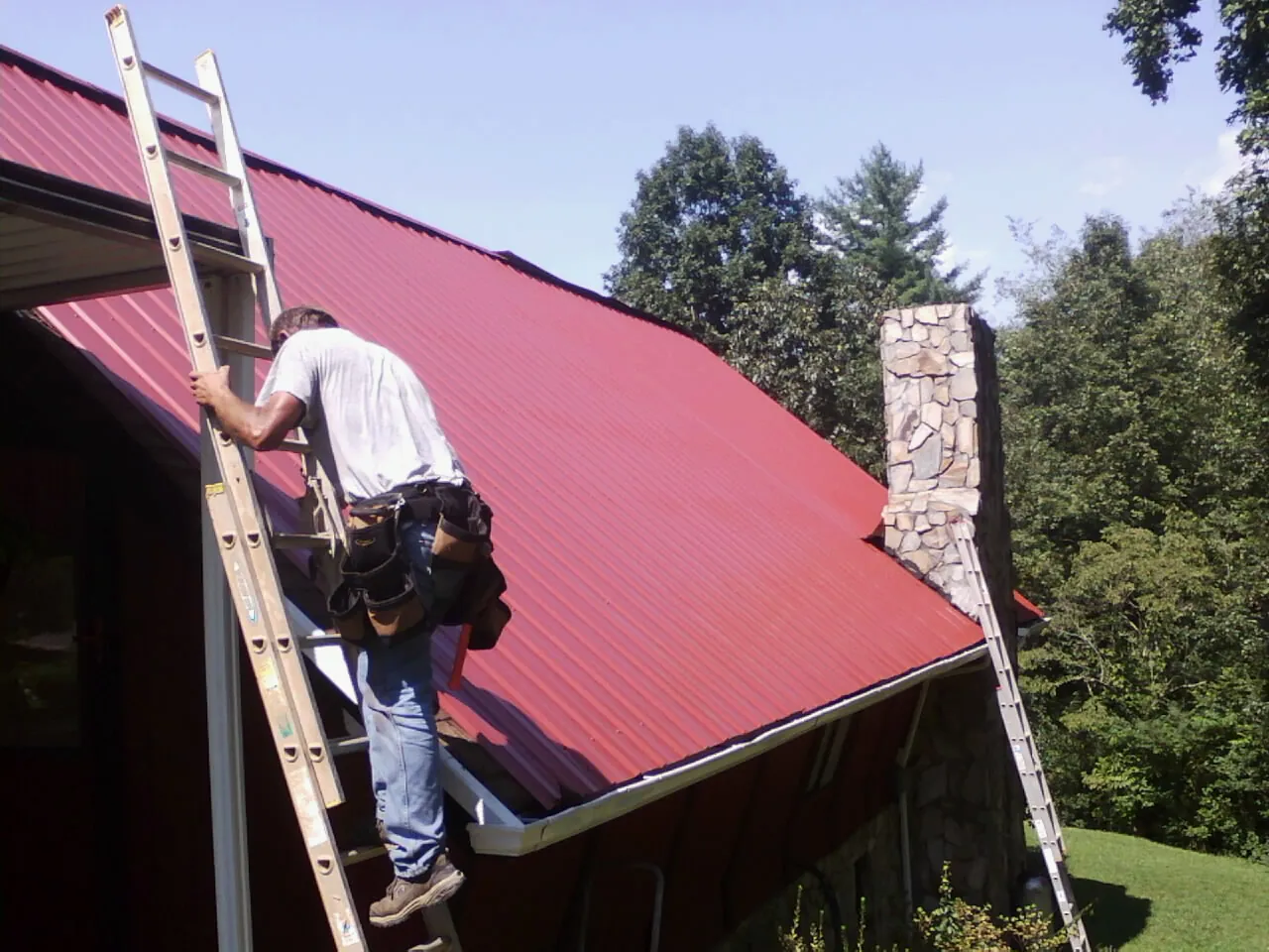 Roofer on a ladder beside a red metal roof and stone chimney — Soffit & Fascia Repair by Roofing Brothers in Sandy Level